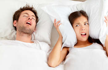 A snoring man sleeping next to a woman covering her ears with the pillow.