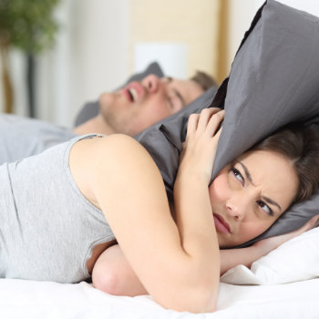 a woman covering her head with a pillow while the man sleeping next to her is snoring