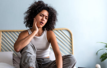 young woman with curly hair sitting on the bed and touching cheek because of toothache