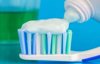 Close-up of toothpaste being dispensed from a tube onto a toothbrush, with a green glass in the background.