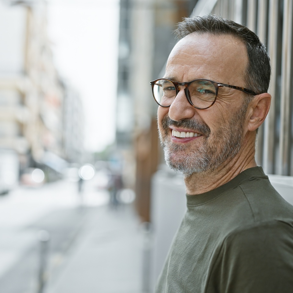 Brimming with joy, middle age man with grey hair stands confidently on city street, his smile beaming happiness as he subtly laughs, his mature expression telling tales of a fulfilled life.