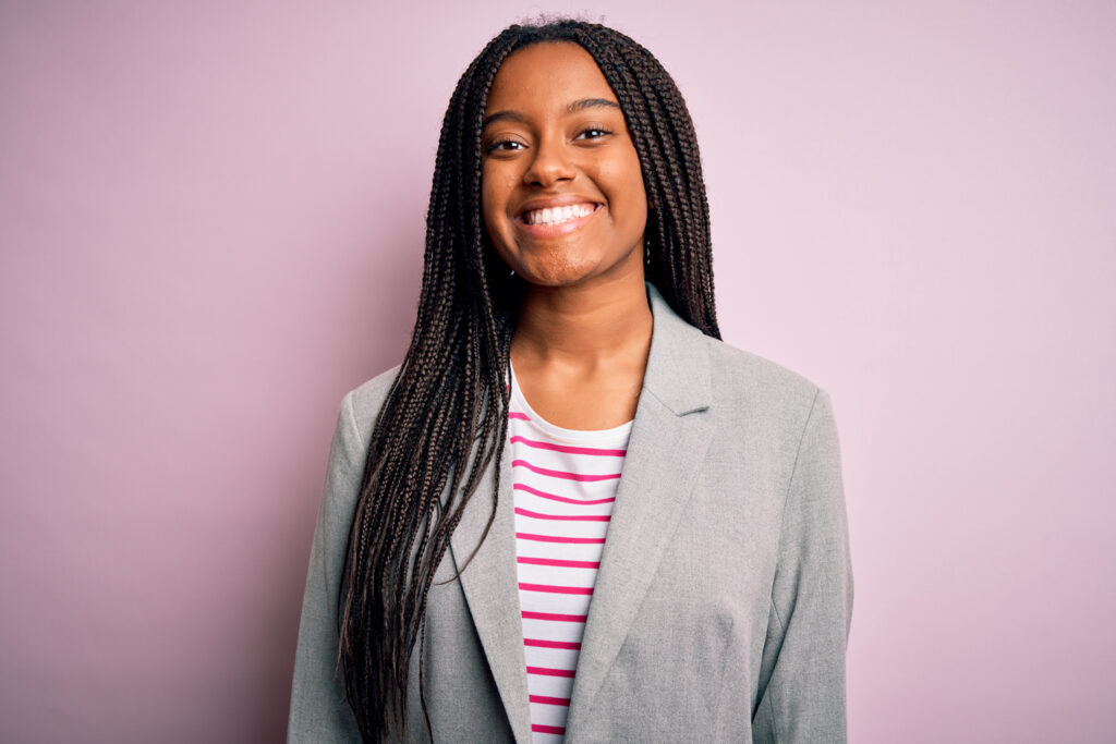 A smiling young woman with long braids wearing a gray blazer and striped shirt standing against a pink background.