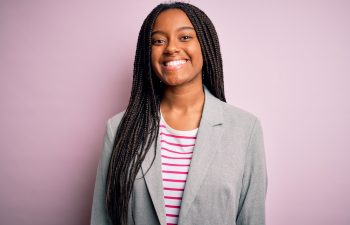 A smiling young woman with long braids wearing a gray blazer and striped shirt standing against a pink background.