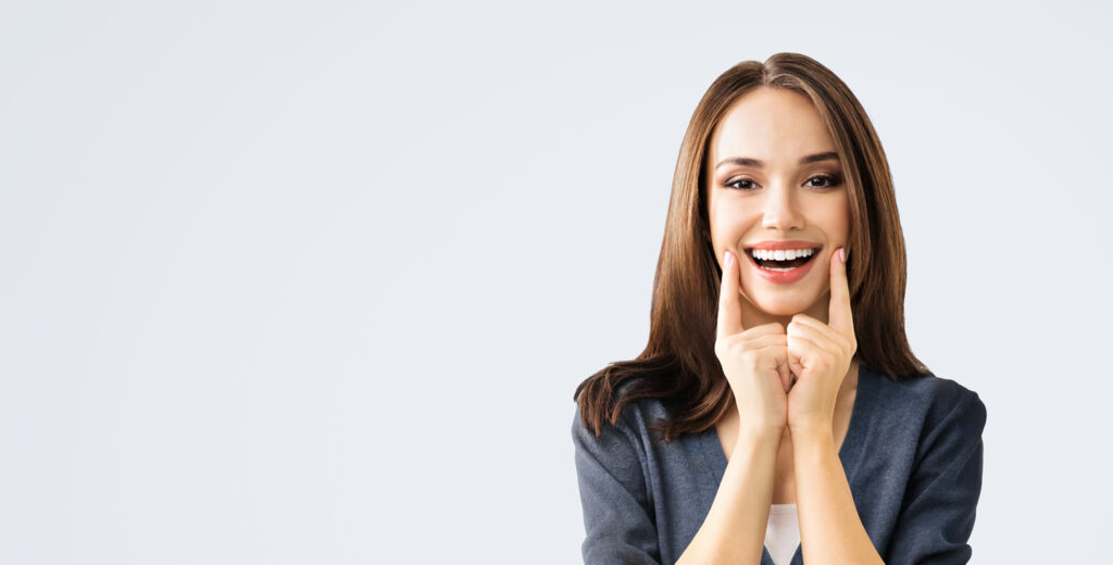 A happy young woman with long, brown hair smiles broadly while pointing to her teeth with both index fingers. She is wearing a dark blazer and is positioned on the right side of the frame against a plain, light gray background, with ample copy space to the left.