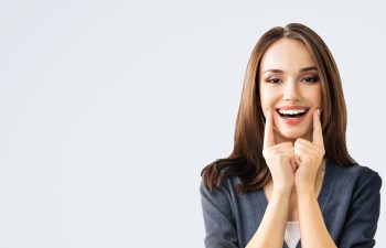 A happy young woman with long, brown hair smiles broadly while pointing to her teeth with both index fingers. She is wearing a dark blazer and is positioned on the right side of the frame against a plain, light gray background, with ample copy space to the left.