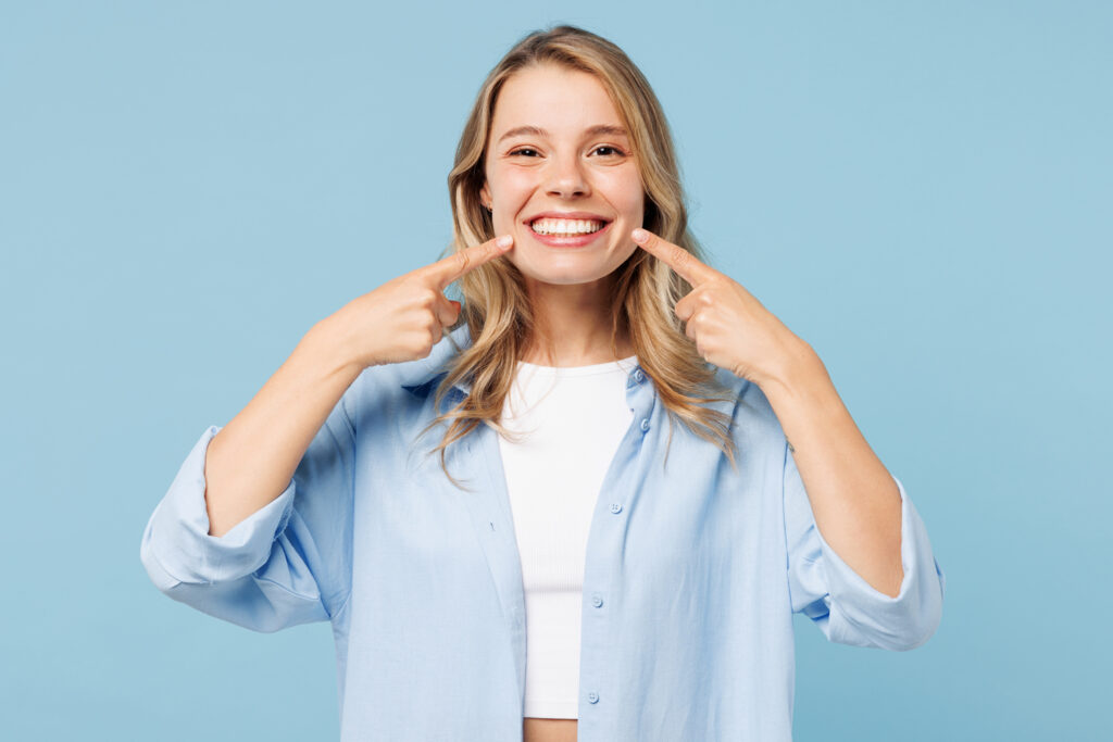 A smiling young woman with blonde hair, wearing a light blue button-up shirt over a white top, points with both index fingers to her bright, healthy smile. The background is solid light blue.
