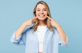 A smiling young woman with blonde hair, wearing a light blue button-up shirt over a white top, points with both index fingers to her bright, healthy smile. The background is solid light blue.