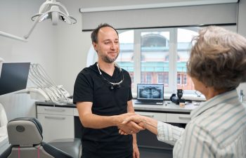 A smiling male dentist wearing black scrubs and dental loupes shakes hands with an older female patient in a modern dental exam room.