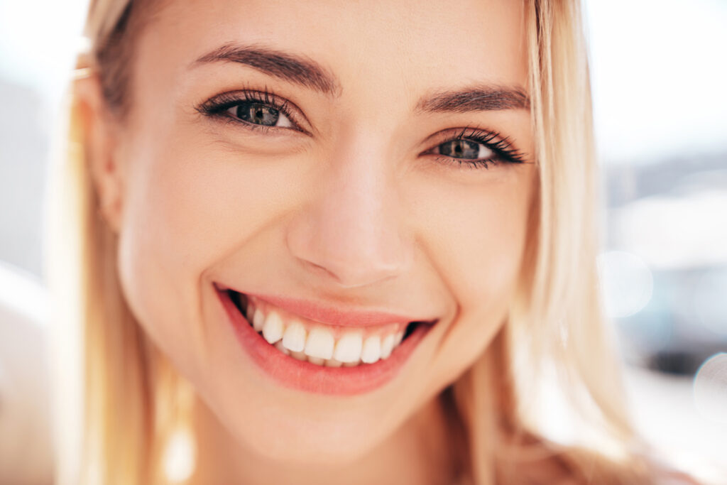 A close-up of a smiling young woman with blonde hair and light-colored eyes, showing a wide, toothy grin at the camera against a brightly blurred background.