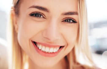 A close-up of a smiling young woman with blonde hair and light-colored eyes, showing a wide, toothy grin at the camera against a brightly blurred background.
