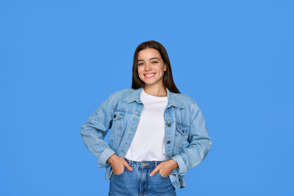 Young woman in a denim jacket and jeans stands smiling with hands in pockets against a solid blue background.