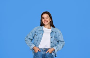 Young woman in a denim jacket and jeans stands smiling with hands in pockets against a solid blue background.