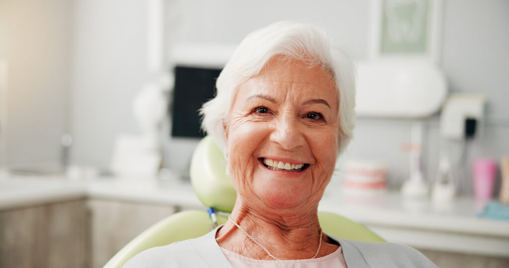 A senior woman with short white hair smiles warmly at the camera while sitting in a dental chair inside a bright dentist's office.