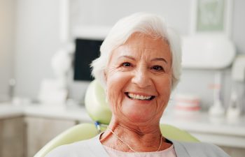 A senior woman with short white hair smiles warmly at the camera while sitting in a dental chair inside a bright dentist's office.