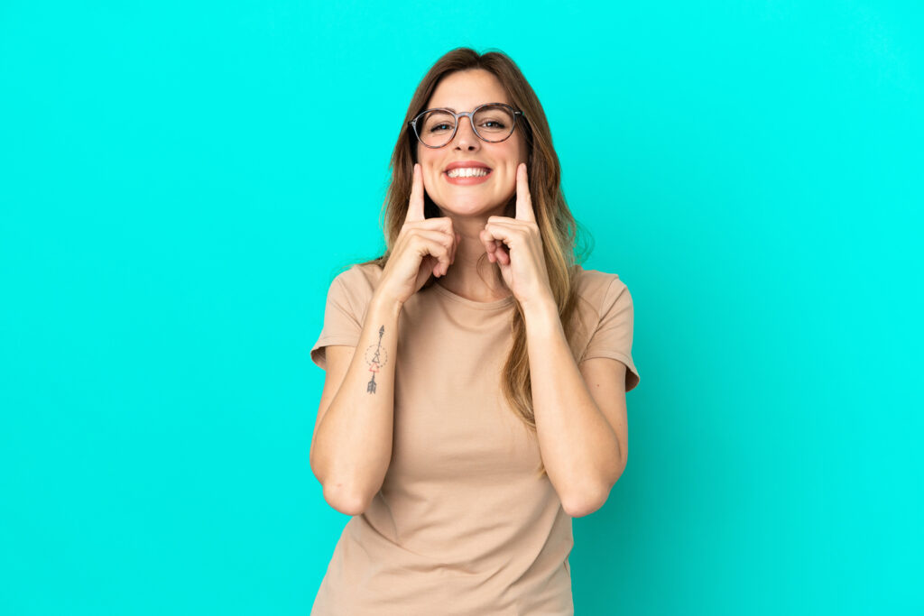 A happy woman with glasses pointing to her cheeks and smile with both index fingers against a bright turquoise background.