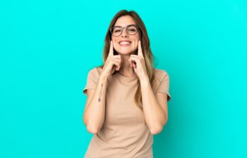 A happy woman with glasses pointing to her cheeks and smile with both index fingers against a bright turquoise background.