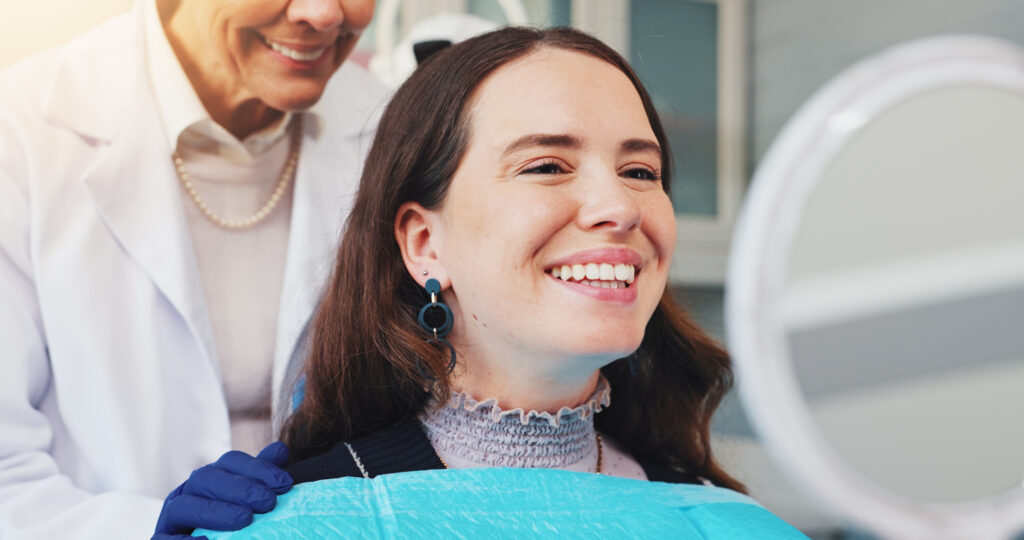 A happy young woman with dark hair sits in a dental chair, wearing a blue bib and looking into a round mirror to check her smile. A dentist in a white coat and blue gloves stands behind her, smiling.