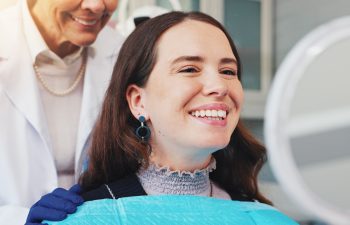 A happy young woman with dark hair sits in a dental chair, wearing a blue bib and looking into a round mirror to check her smile. A dentist in a white coat and blue gloves stands behind her, smiling.