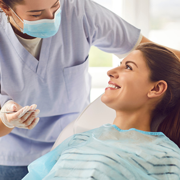 A dentist wearing gloves and a mask talks to a smiling female patient who is sitting in a dental chair with a protective bib.