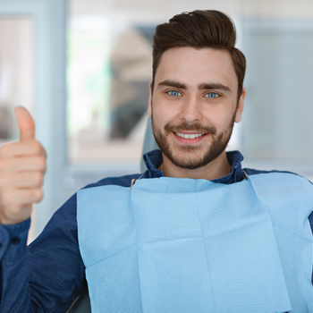 A man sitting in a dental chair wearing a bib gives a thumbs up and smiles at the camera.