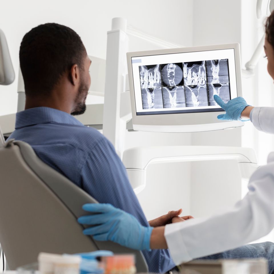A dentist shows a dental X-ray on a monitor to a patient sitting in a dental chair.