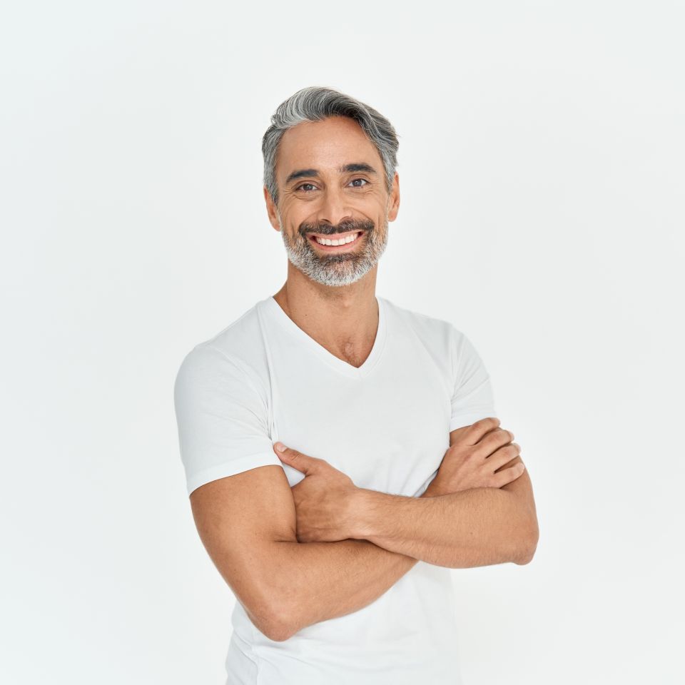 Man with gray hair and beard wearing a white t-shirt stands against a plain white background, smiling with arms crossed.