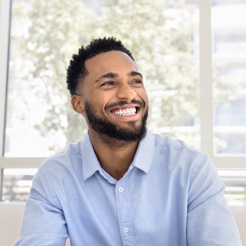 Cheerful attractive young Black man in pale blue shirt looking away with perfect toothy smile in deep positive thoughts, laughing, showing white teeth, promoting dental care