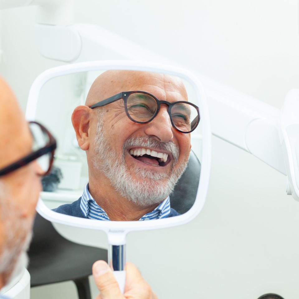 Happy senior patient admiring his perfect smile after a dental implant treatment, holding a mirror in his hand at the dental clinic